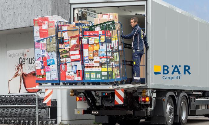 Lkw-Fahrer lädt mehrere Lebensmittelrollbehälter mit Hilfe des Bär Cargolifts vor dem Lebensmittelgeschäft aus dem Lkw 